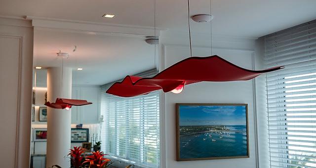 Luce Volante in red hanging in a white walled room with a painting hung on one of the walls and windows on the other side