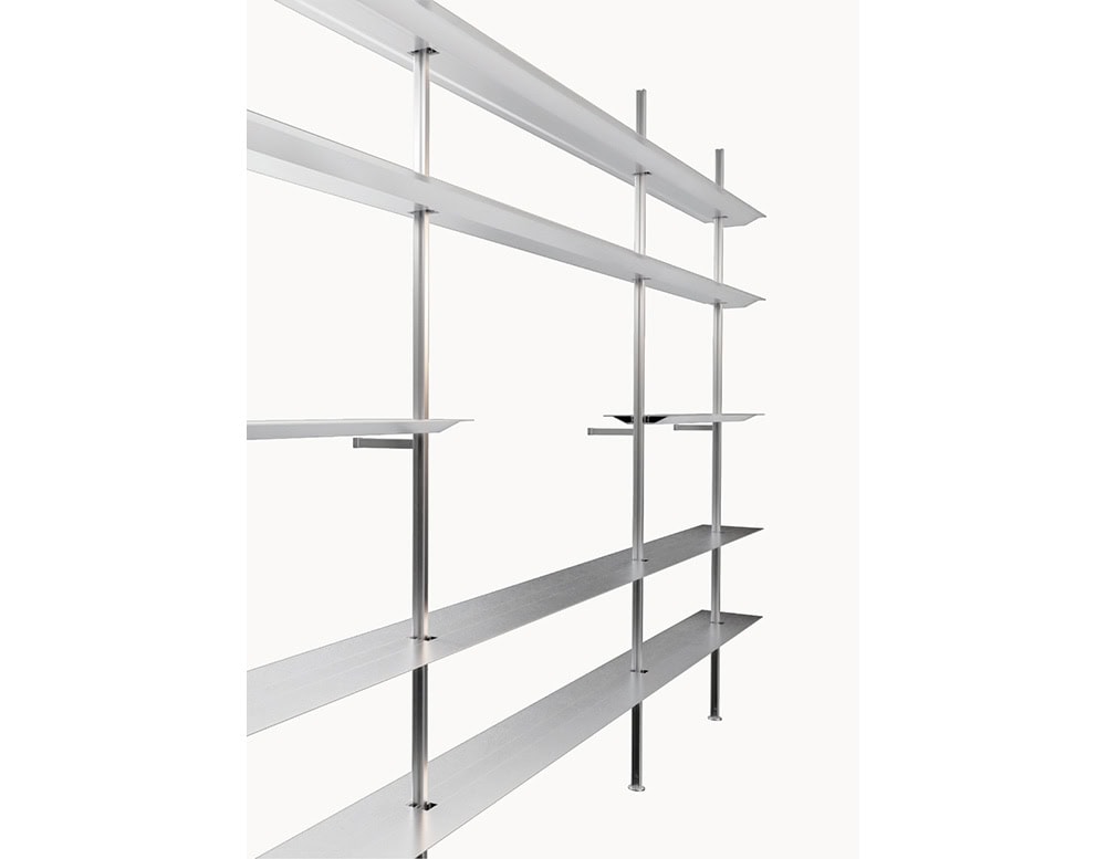 Modular shelving unit with slim vertical posts and wide, brushed metal shelves. Its open, linear design offers a light and architectural presence.