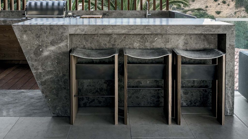 Three Rio Stools, structure and two legs in natural wood with central crosspiece covered in brass; seat in gray leather in a kitchen.