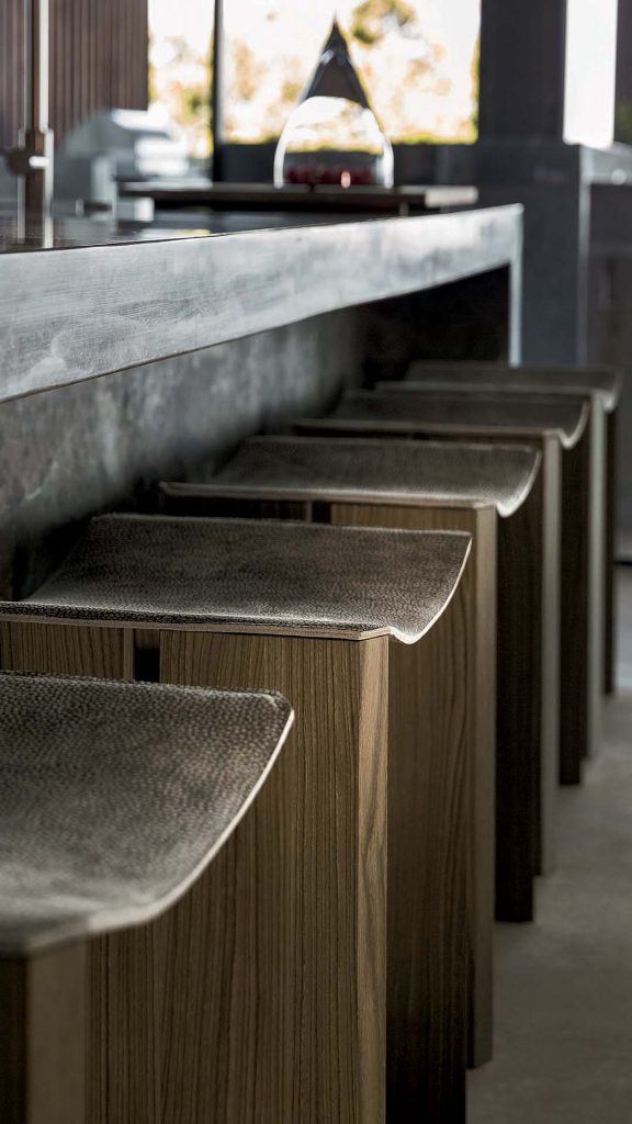 Closeup of four Rio Stools, structure and two legs in natural wood with central crosspiece covered in brass and seat made of gray leather.