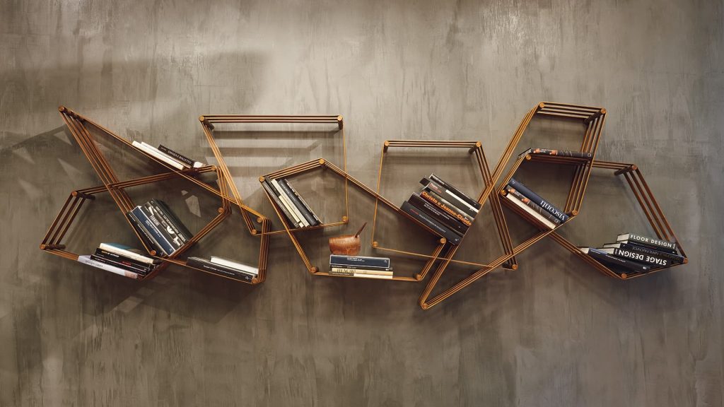Tangram bookcase made of irregular polygons joined together. Structure in natural burnished brass attached to a gray wall.