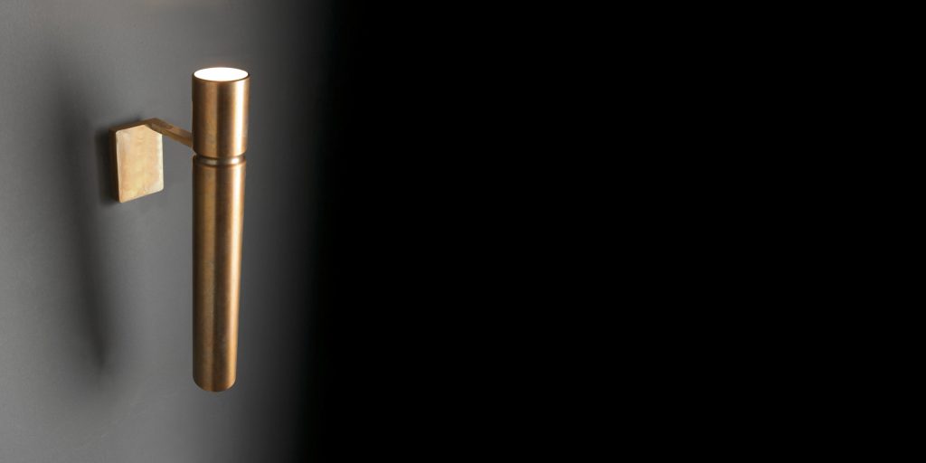 Ceiling Tubular Light with a cylindrical shape in bronze attached to a black wall with a bronze support.