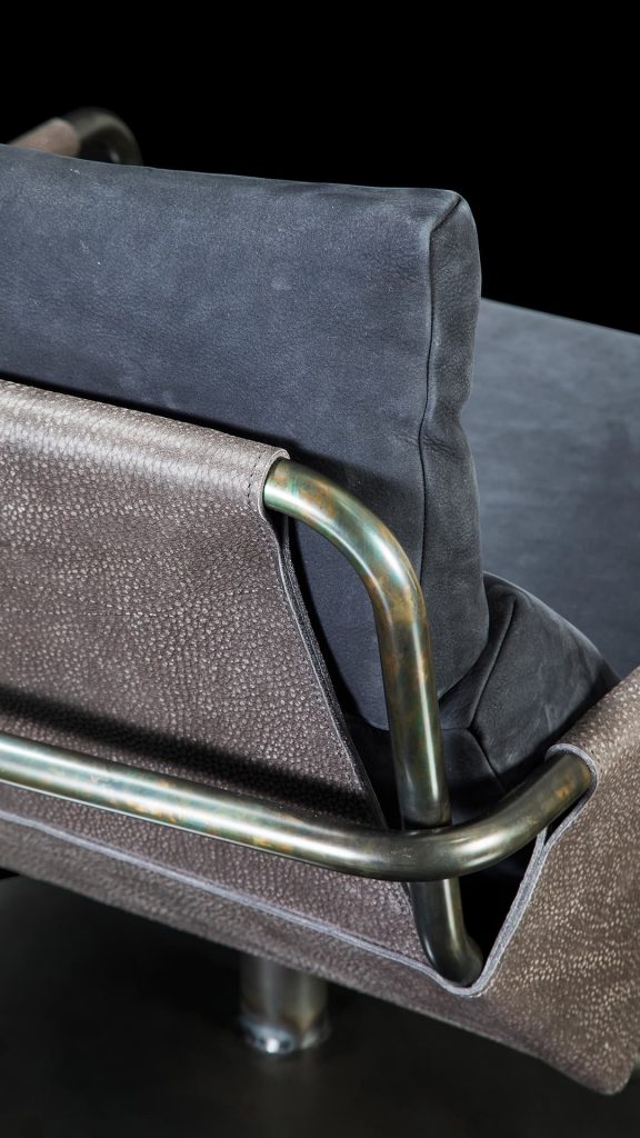 Closeup of Voyage armchair, curved bronze steel structure with brown leather, gray leather cushion upholstery, central black steel leg on a black background.