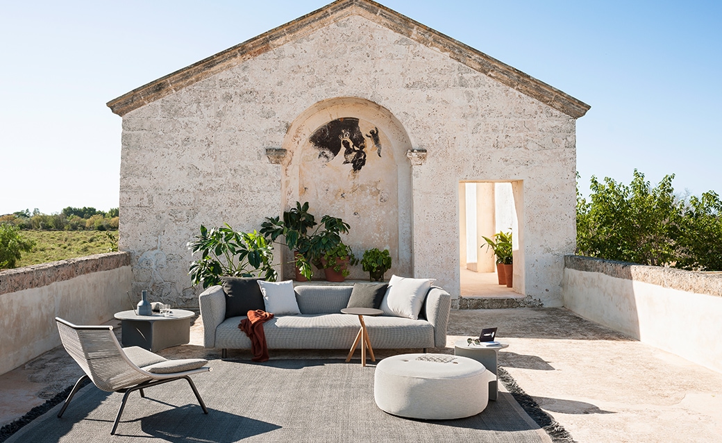 Outdoor lounge area with a modern sofa, pouf, and lounge chairs set against a rustic stone building with lush greenery