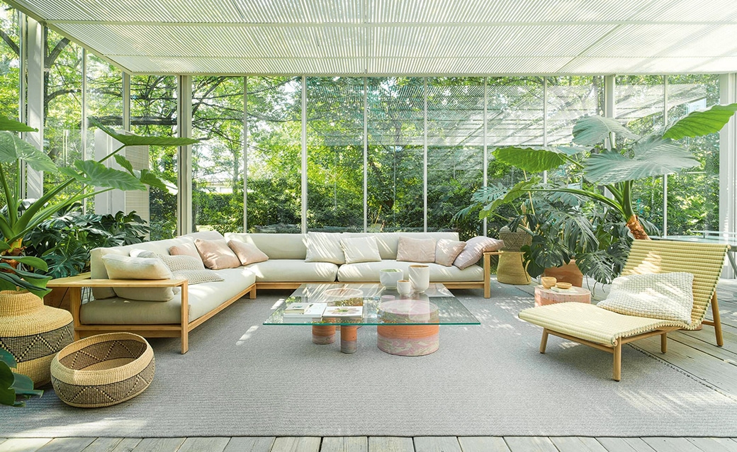 A bright and airy indoor-outdoor living space featuring the Frei sectional sofa in natural wood and rattan with beige cushions, paired with a glass coffee table, surrounded by lush greenery and floor-to-ceiling windows.