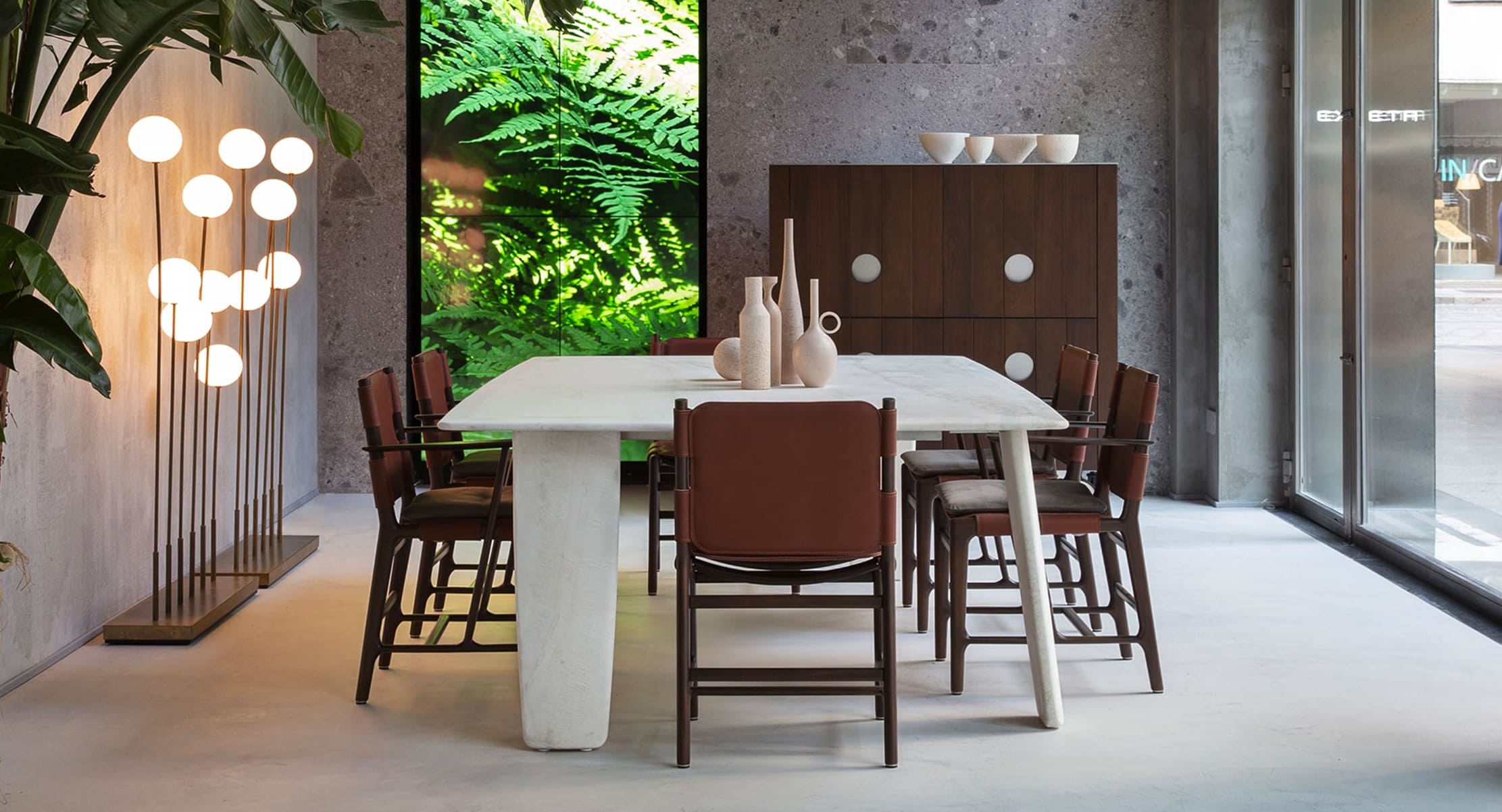 A modern dining area featuring the Manto dining table with a sculptural white base, surrounded by wooden chairs with leather seats, set against a lush green backdrop.