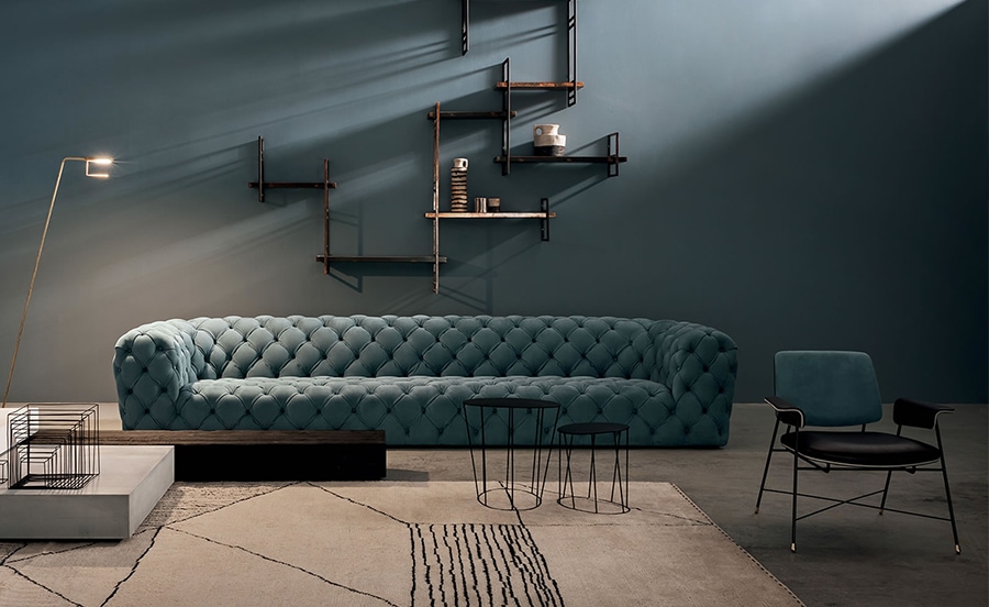 A deep blue tufted Chester Moon sofa with a luxurious quilted design, set against a dark moody interior with floating shelves and contemporary decor