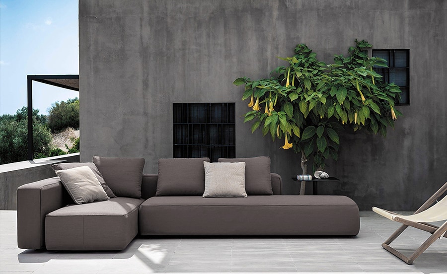 Dandy – A stylish outdoor modular sofa with deep brown cushions, set against a modern concrete wall with a lush green plant adding a touch of nature