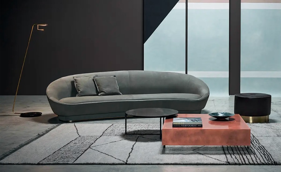 A sleek, modern curved sofa in muted gray with a smooth, sculptural design, set in a sophisticated interior featuring geometric rugs, a minimalist black wall, and a glass window