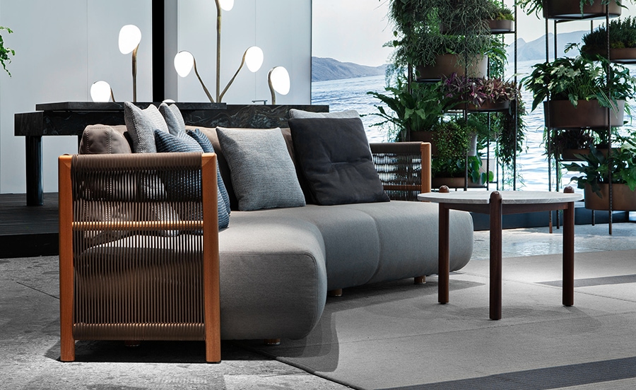 Levante – A sophisticated outdoor sofa with plush gray cushions, wooden armrests, and a sleek modern frame, set against a serene backdrop of greenery and water