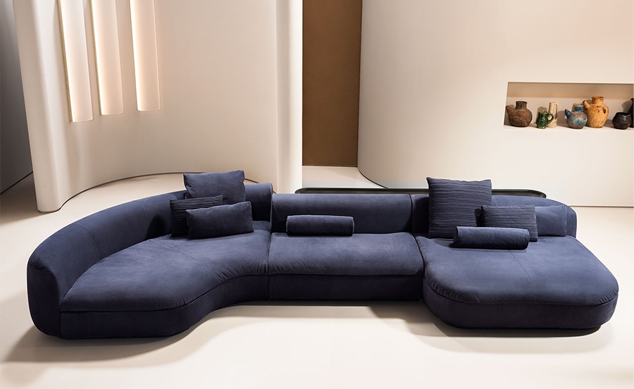 A contemporary modular sofa in deep navy blue with a curved, ergonomic design, plush cushions, and adjustable headrests, set in a minimalist interior with warm lighting