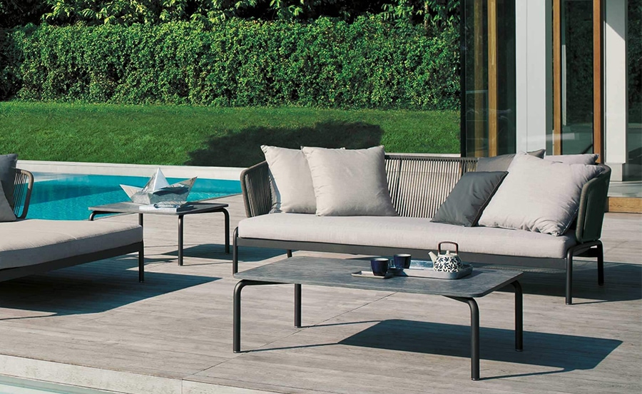 Spool – A modern outdoor sofa with a sleek metal frame and light beige cushions, set beside a swimming pool with lush green landscaping in the background