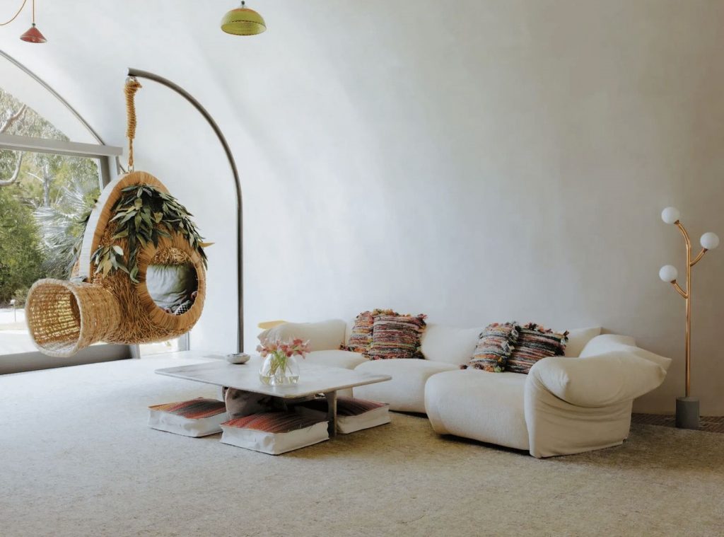 A modern living space with a curved white ceiling, a cozy off-white sofa, a hanging wicker chair with lush greenery, and a low coffee table on a neutral rug