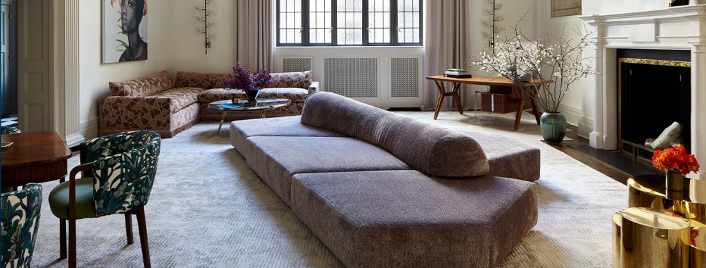 A modern living room featuring a large, plush, dark gray sofa with a cylindrical cushion, set against a backdrop of tall windows with natural light streaming in, soft curtains, and classic radiator covers