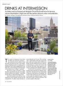 A man stands on a stylish rooftop terrace with lush greenery, overlooking a city skyline with tall buildings in the background.