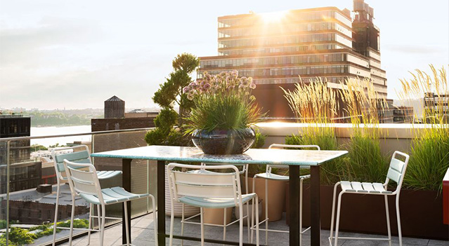 A stylish rooftop dining area bathed in warm sunlight, featuring a sleek modern table, minimalist white chairs, and lush greenery enhancing the urban ambiance