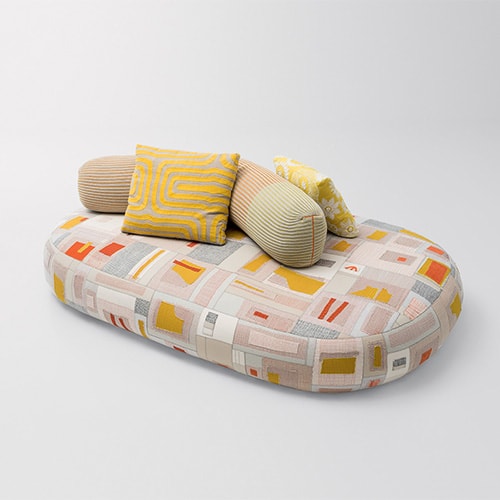 Indoor seating platform of fabric with geometric figures in different warm and gray colors, with a backrest composed of a cylindrical element with the fabric of green and orange lines, and cushions of yellow textures