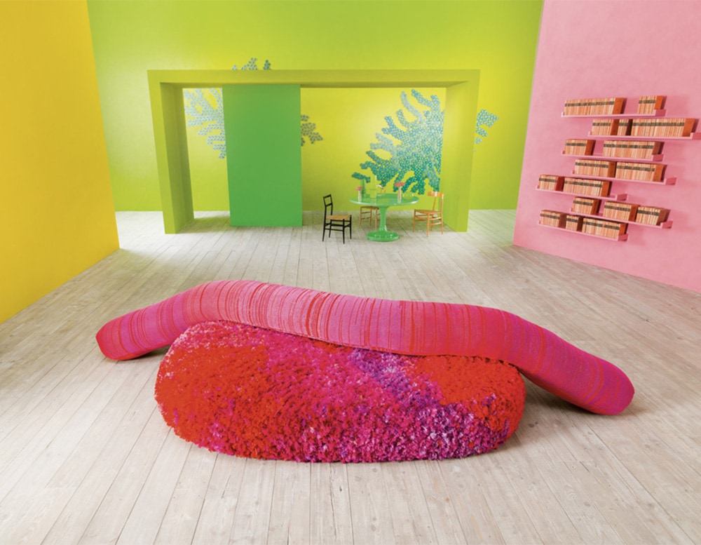 Indoor seating platform covered with fuchsia and violet fabric with a pink backrest composed of a long cylindrical element in a room with green background