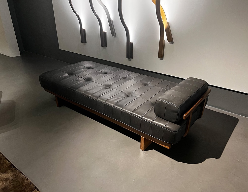 Side view of a daybed of black leather atop a frame made of filigree wood slats of brown color