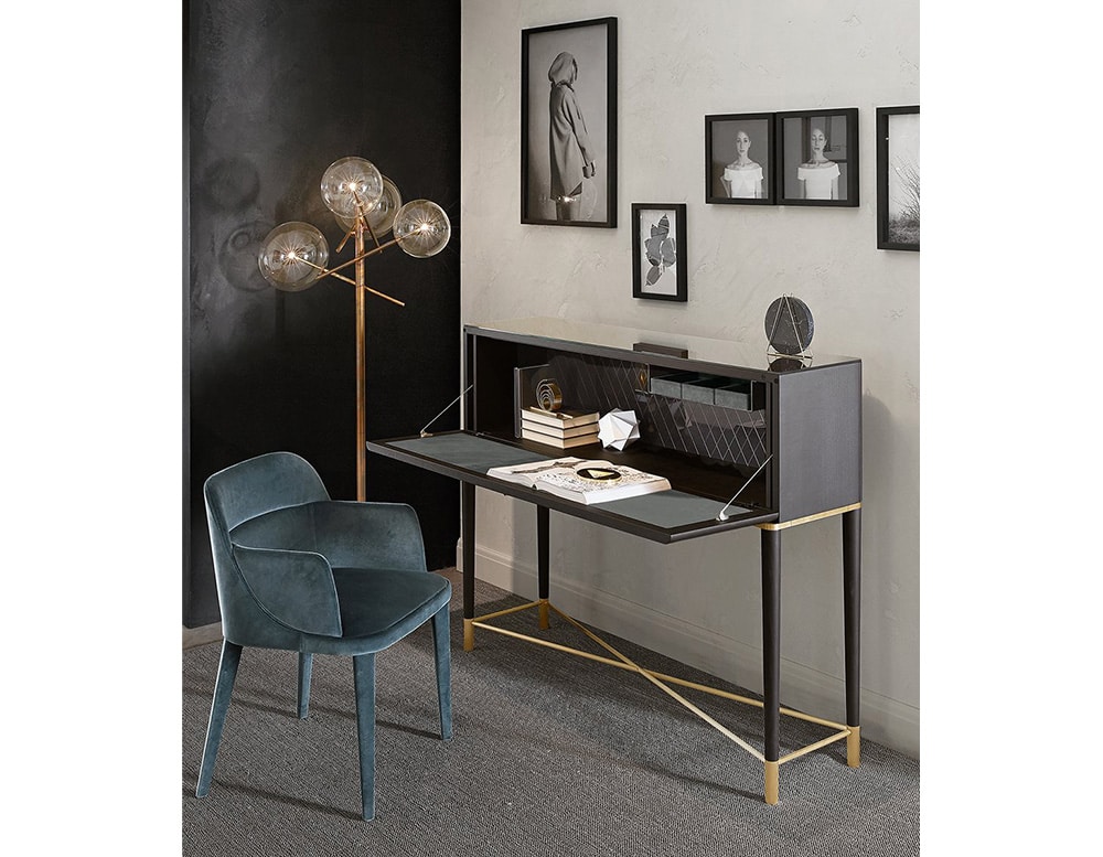 Writing desk with flap door in black wood with borders of satin brass lacquered metal parts and bright licorice painted glass top and inside back with a rhomboidal engraved pattern on the inside glass back beside a blue chair