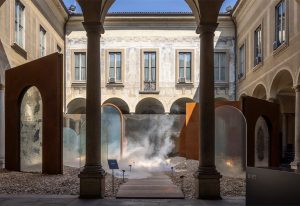 A historic courtyard framed by stone columns, featuring mist rising from the ground alongside modern rust-colored and glass structures