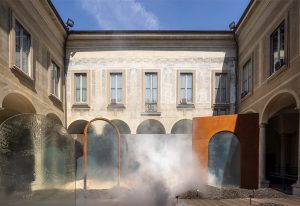 A historic courtyard with arched windows and a misty installation, featuring modern rust-colored and glass structures blending old and new