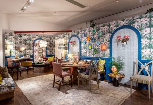 A brightly decorated room features a wooden dining table with four chairs, a patterned rug, and walls covered in colorful, whimsical wallpaper with arched doorways painted in blue. The room has a playful, eclectic aesthetic.