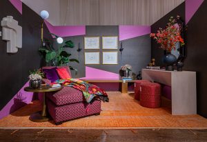 A vibrant interior with geometric pink and brown walls, a plush red armchair with a colorful throw, a modern desk, and warm lighting