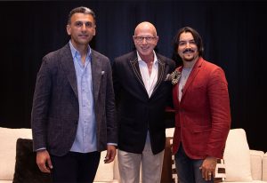 Three men standing side by side in formal attire, posing for a photograph with a dark curtain backdrop and white seating behind them