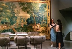 A sophisticated interior setting with elegant dining chairs and a large classical tapestry, as two women engage in conversation