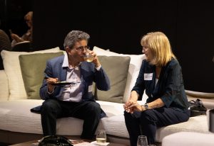 A man in a blue suit and a woman in a black outfit sit on a white couch, engaged in conversation while enjoying food and drinks