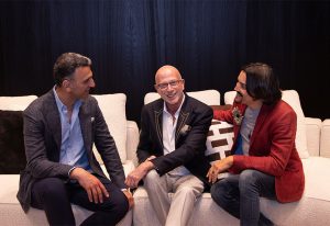 Three men sitting on a white couch in conversation, dressed in formal attire, with one man in the center smiling and engaging warmly