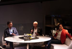 Three individuals seated around a round table in a dimly lit event space, engaged in a discussion with microphones and drinks present
