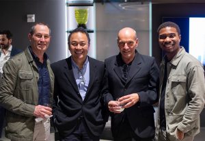 A group of four men dressed in stylish attire pose together at a social design event, each holding a drink and smiling at the camera