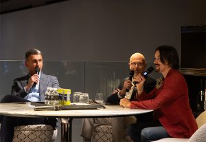 Three individuals engaged in a panel discussion at a modern event space, seated around a table with a microphone and drinks