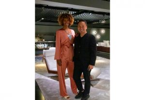 Two elegantly dressed individuals stand together in a modern, well-lit showroom with luxurious furniture and ambient lighting in the background