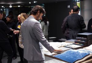 A man in a gray suit examines fabric and design samples on a table, surrounded by other attendees at a stylish design event