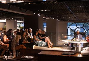 An audience attentively listens during an indoor event in a dimly lit, modern space with stylish furnishings and warm lighting