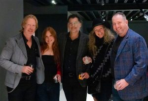 A group of five elegantly dressed people pose together at a social event, holding drinks and smiling in a stylish, dimly lit setting