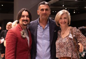 Three people pose together, smiling at the camera in a warmly lit indoor space, dressed in stylish and semi-formal attire