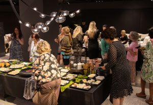 A lively gathering with elegantly dressed guests enjoying a buffet spread in a dimly lit space with modern pendant lighting overhead