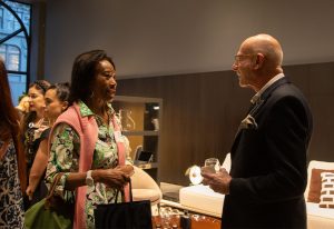A woman in a floral dress and a man in a suit hold drinks while engaged in conversation in a dimly lit, elegant setting