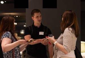 A man in a black shirt with a name tag holds a plate of food while conversing with two women in a warmly lit indoor setting