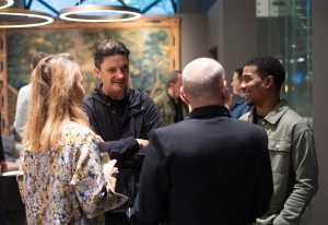 A group of elegantly dressed individuals engaged in conversation in a sophisticated setting with an artistic backdrop and warm lighting