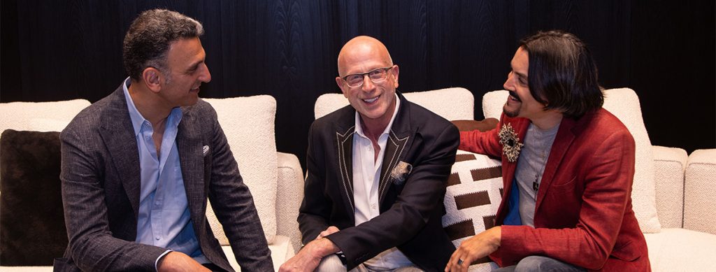 A smiling bald man in a dark blazer and light-colored shirt sits on a stylish couch, engaged in conversation, with elegant decor and plush cushions in the background.