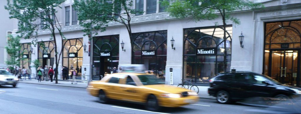 A blurred yellow taxi drives past a high-end Minotti showroom with large glass windows and elegant exterior lighting in an urban setting