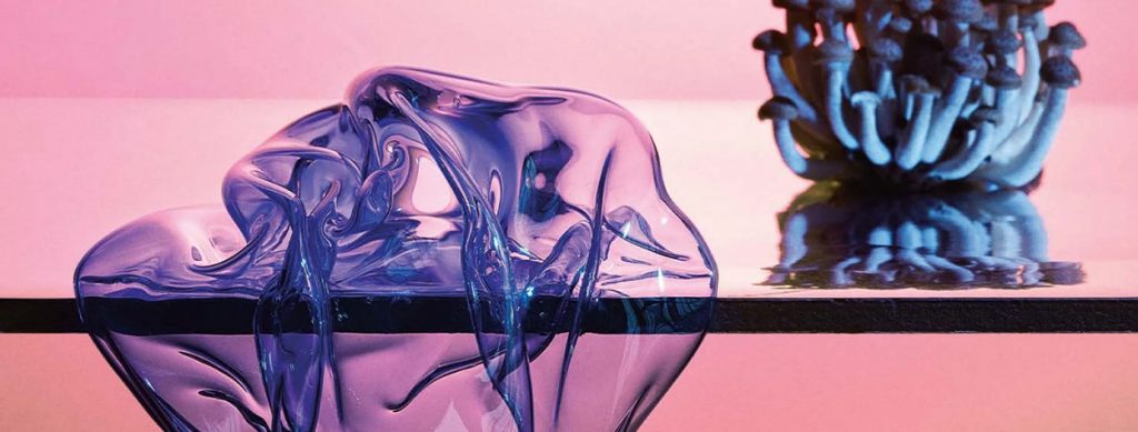 A close-up of an artistic glass sculpture in shades of blue and purple, resting on a reflective surface against a soft pink gradient background