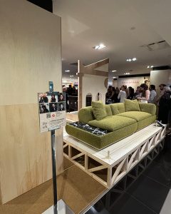 A green sofa is displayed on a raised platform in a public space, possibly a showroom or event, with people visible in the background
