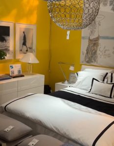 A bold yellow wall sets a radiant backdrop for crisp black-and-white bedding and soft-gray pillows. Sleek white furniture, soft lighting, and modern art create a cozy yet striking bedroom with an editorial edge.