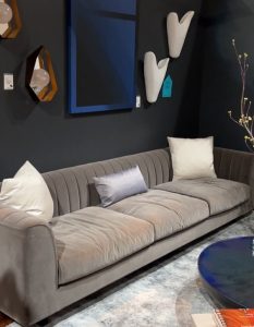 A deep-gray velvet sofa anchors this moody corner, flanked by sculptural sconces, geometric mirrors, and cobalt blue wall art. Plush pillows soften the scene, while subtle lighting adds drama to the charcoal-painted backdrop.
